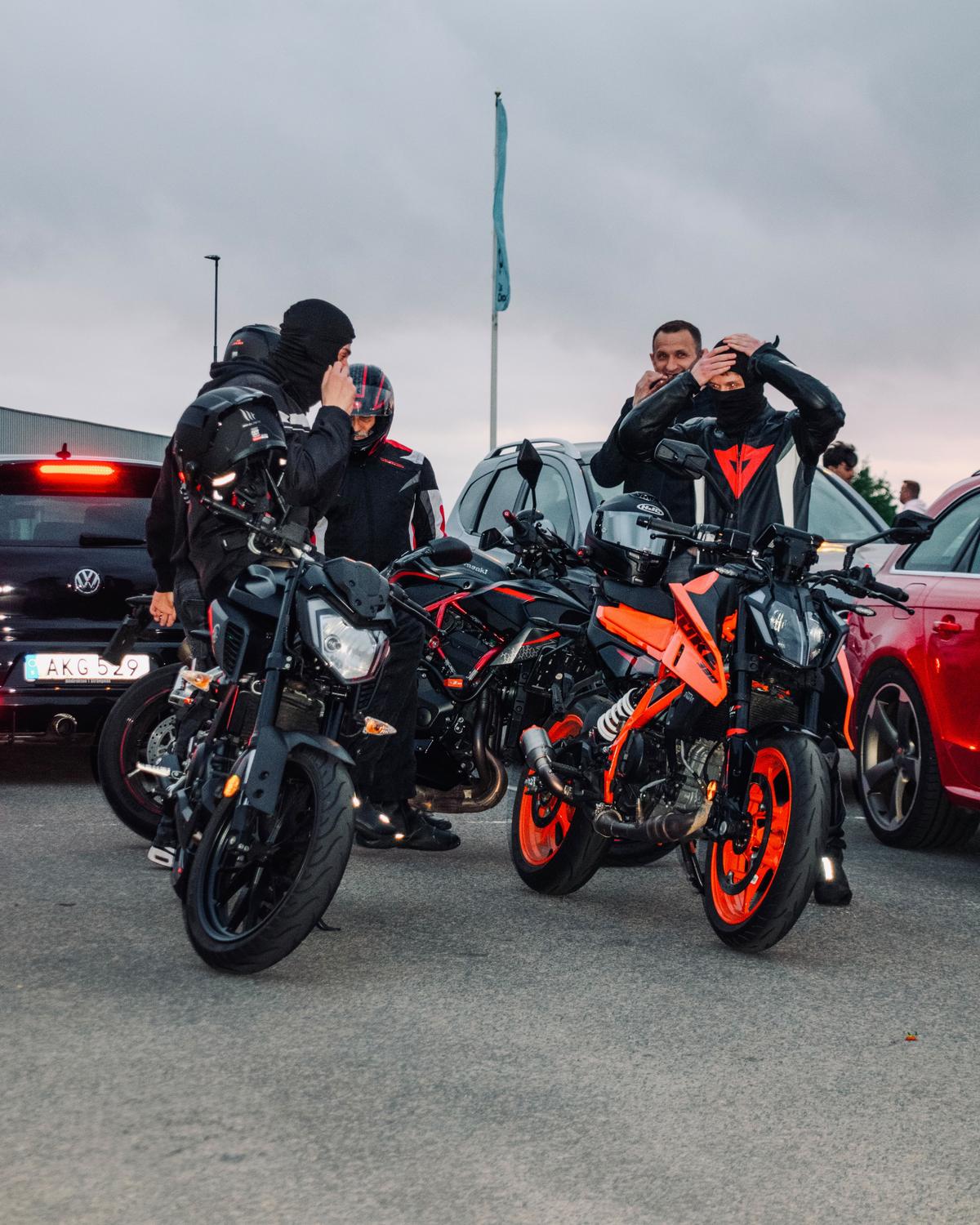Fyra motorcyklister i skinnställ tar på sig hjälmar vid sina motorcyklar på en parkering i skymningen